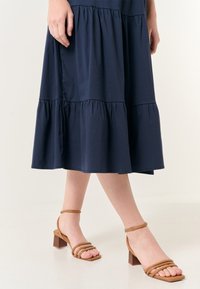 Marineblaues schichtweise Kleid mit einem Rüschenrand, kombiniert mit tanfarbenen Sandalen mit Riemchen, die Blockabsätze und ein quadratisches Zehengestaltung aufweisen.