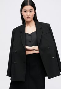 Μαύρο διπλό σακάκι με υφή, με μεγάλα κουμπιά και δομημένη σιλουέτα. Φορεμένο πάνω από ένα εφαρμοστό σκούρο γκρι τοπ.