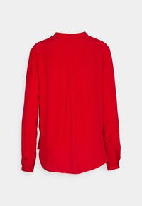 Blouse rouge à manches longues avec une coupe ample, un ourlet arrondi, un pli au dos et des poignets boutonnés, présentée sur un fond blanc.