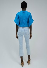 Blusa de lino azul con mangas anchas, combinada con unos vaqueros recortados de un azul claro y tacones marrones. La parte posterior tiene un detalle de pliegue.