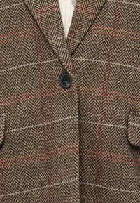 Veste en tweed brun chevron avec carreaux rouge et bleu clair, un bouton noir unique et poches avant à rabats.