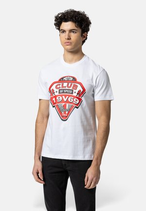 Jonge man draagt een wit T-shirt met een rood-zwarte "Club of Speed 19V69 Racer Italia" print, gecombineerd met een zwarte broek.