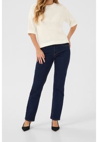 Pull crème en tricot à manches courtes, associé à un jean droit bleu foncé et des talons noirs à bout pointu, posé sur un fond clair.