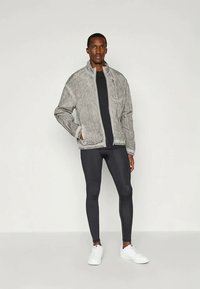 Pilkas užtrauktuku užsegamas švarkas su tekstūruotu audiniu, juodos prigludusios Leggingos ir baltos sportbačiai. Modelis stovi neutralioje fone.