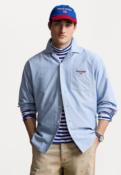 Light blue button-up shirt with a pocket, layered over a blue and white striped turtleneck. Blue and red cap with "POLO SPORT" logo. Tan pants with paint stains.