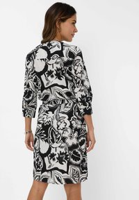 Schwarzes und weißes Kleid mit Blumenmuster, dreiviertelÄrmeln, einer taillierten Taille und geradem Schnitt. Der Stoff wirkt leicht und strukturiert.