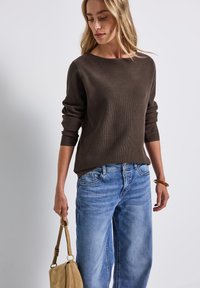 Maglione marrone a coste con scollatura ampia, abbinato a jeans a gamba larga di un blu chiaro. Una borsa beige è tenuta in una mano; texture morbida e vestibilità casual.