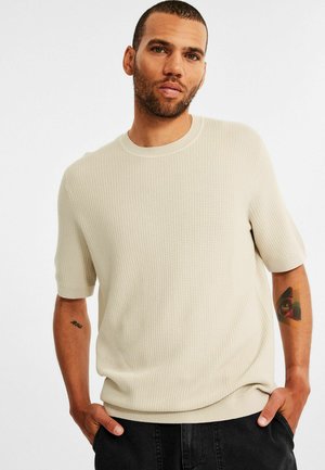 Lys beige kortærmet sweater med et tekstureret, ribbet mønster. Har en rund halsudskæring og en afslappet pasform. Sorte jeans og en arm tatovering synlig.