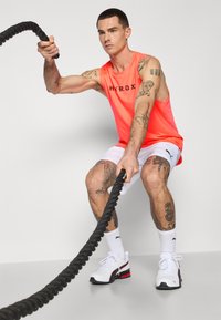 Neon orange ærmeløs tank top med "HYROX" logo, hvide shorts, sort battle rope og hvide sneakers med røde og sorte detaljer.
