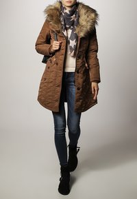 Brun quiltet parka med pelsforet hætte, parret med et stjernemønstret tørklæde, let sweater, blå jeans og sorte ankelstøvler.