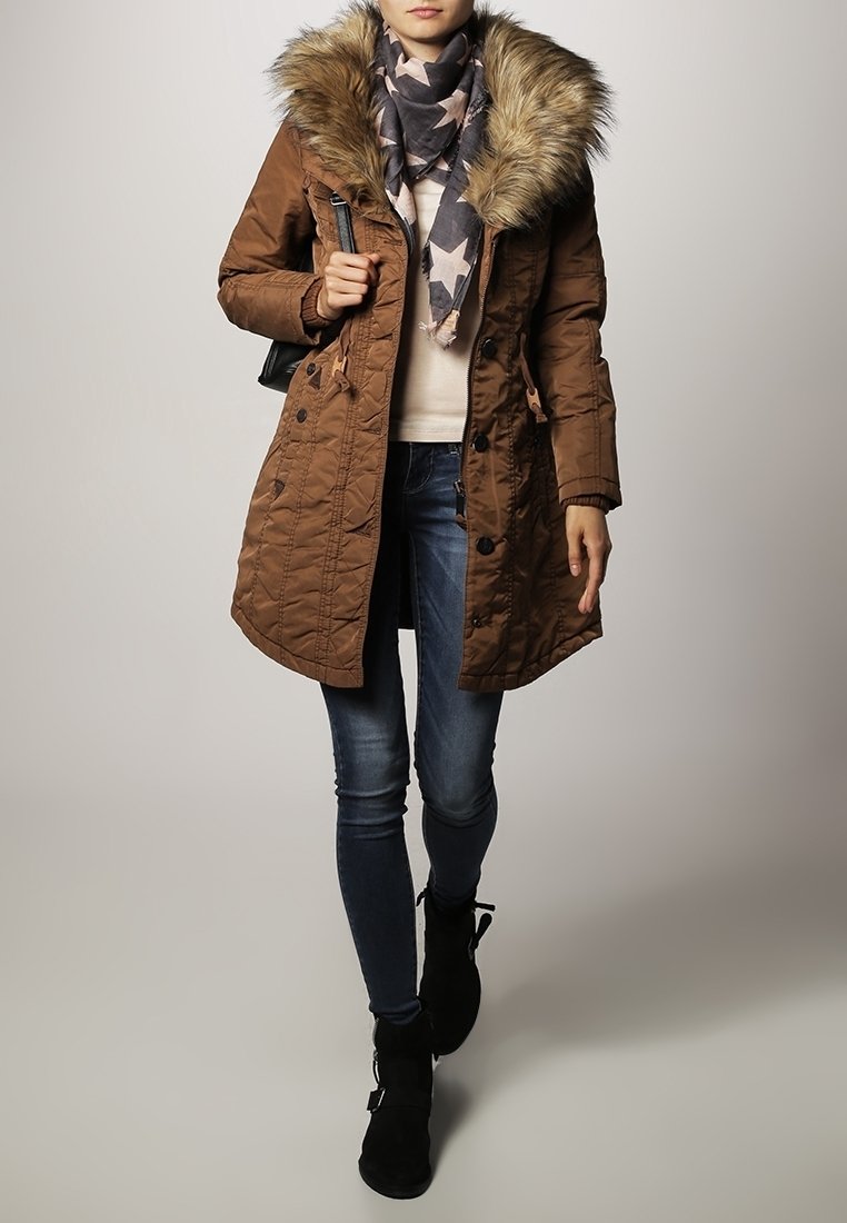 Brun quiltet parka med pelsforet hætte, parret med et stjernemønstret tørklæde, let sweater, blå jeans og sorte ankelstøvler.