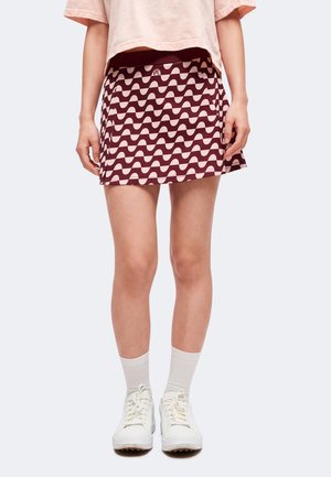 Femme portant une mini-jupe à motifs bordeaux et beige, un crop top rose pâle, des chaussettes blanches et des baskets blanches, debout devant un fond uni.