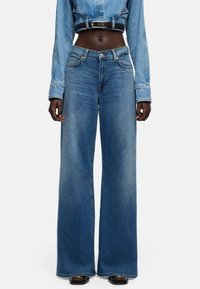 Szerokie, niebieskie dżinsy z denimu o wyblakłym wykończeniu, z wysokim stanem i pięciokieszeniowym krojem, stylizowane z krótką kurtką dżinsową i paskiem.