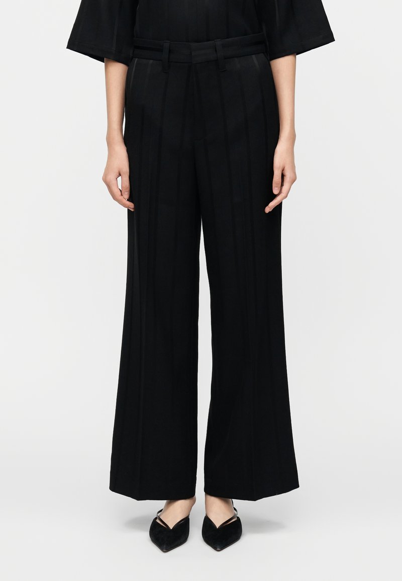 Plačios juodos kelnės su vertikaliomis siaurėjančiomis juostelėmis, turi lygią priekinę dalį, diržo kilpas ir pasiūtas pagal figūrą. Derinamos su juodais, smailiais batais.