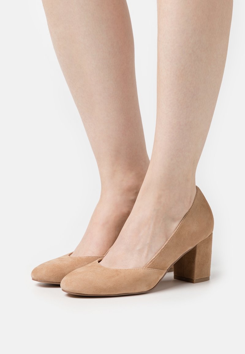 Pieds de femme portant des escarpins en daim beige à talons carrés avec des bouts arrondis sur un fond blanc.