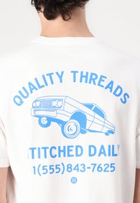 Person wearing white t-shirt with blue graphic of a lowrider car and text "Quality Threads Stitched Daily" and a phone number on the back.