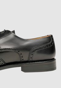 Chaussure de ville en cuir noir avec un bout arrondi, des détails brogues et une texture lisse. Semelle en caoutchouc épaisse avec un motif de traction subtil.