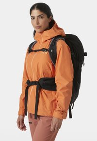 Chaqueta impermeable naranja con capucha ajustable, cinturón negro en la cintura y bolsillos laterales, llevada sobre pantalones marrones. Mochila visible en la espalda.