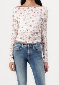 Mujer que lleva una blusa blanca de encaje con hombros descubiertos y mangas largas, con patrones florales rojos, y jeans azul de lavado medio.