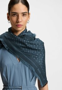 Dark blue scarf with a geometric pattern and fringed edges, draped over a light blue garment, featuring the brand name "BOSS" in the design.