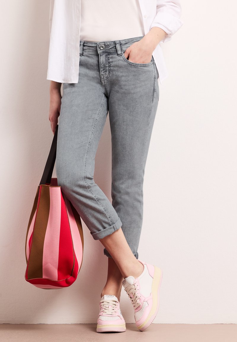 Personne portant un jean gris retroussé, des baskets roses et blanches, une chemise blanche, tenant un sac fourre-tout rayé rouge, rose et marron sur le côté.