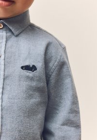 Camicia azzurro chiaro con bottoni, realizzata in tessuto strutturato, con un piccolo ricamo nero di un'auto sulla parte superiore del petto.