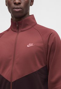 Άνδρας με μπουφάν Nike σε βυσσινί και σκούρο κόκκινο με φερμουάρ, ψηλό γιακά και ροζ λογότυπο Nike στο στήθος, σε απλό φόντο.