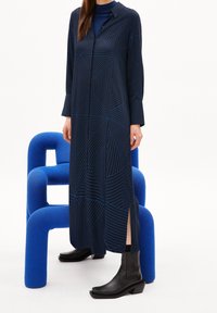 Kvinna klädd i en lång marinblå klänning med geometriska linjer, svarta stövlar, stående framför en blå texturerad modern stol mot vit bakgrund.