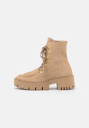 Lichtbruine suède enkellaars met een chunky zool, gouden veterhaken, zijrits en ronde neus op een witte achtergrond.