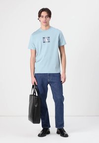 Lys blå bomulls t-skjorte med et svart og hvitt grafisk logo, kombinert med mørkeblå jeans og en svart lær shopper.