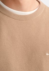 Pull en tricot beige avec un motif texturé, doté d'un col rond et d'un logo discret sur le devant. Doublure intérieure lisse visible.