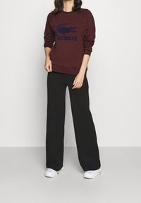 Sweat-shirt bordeaux avec logo bleu marine et poignets côtelés, associé à un pantalon large noir et des baskets blanches. Tissu lisse, coupe décontractée.