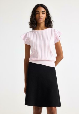 Kvinde med krøllet hår iført en lyserød ribbet top med flæseærmer og en knælængde sort nederdel, stående foran en ensfarvet baggrund.