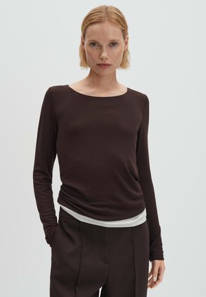 Braunfarbenes Langarmoberteil mit Rundhalsausschnitt, aus glattem Stoff, kombiniert mit einer hellen Basisschicht, die am Saum sichtbar ist.