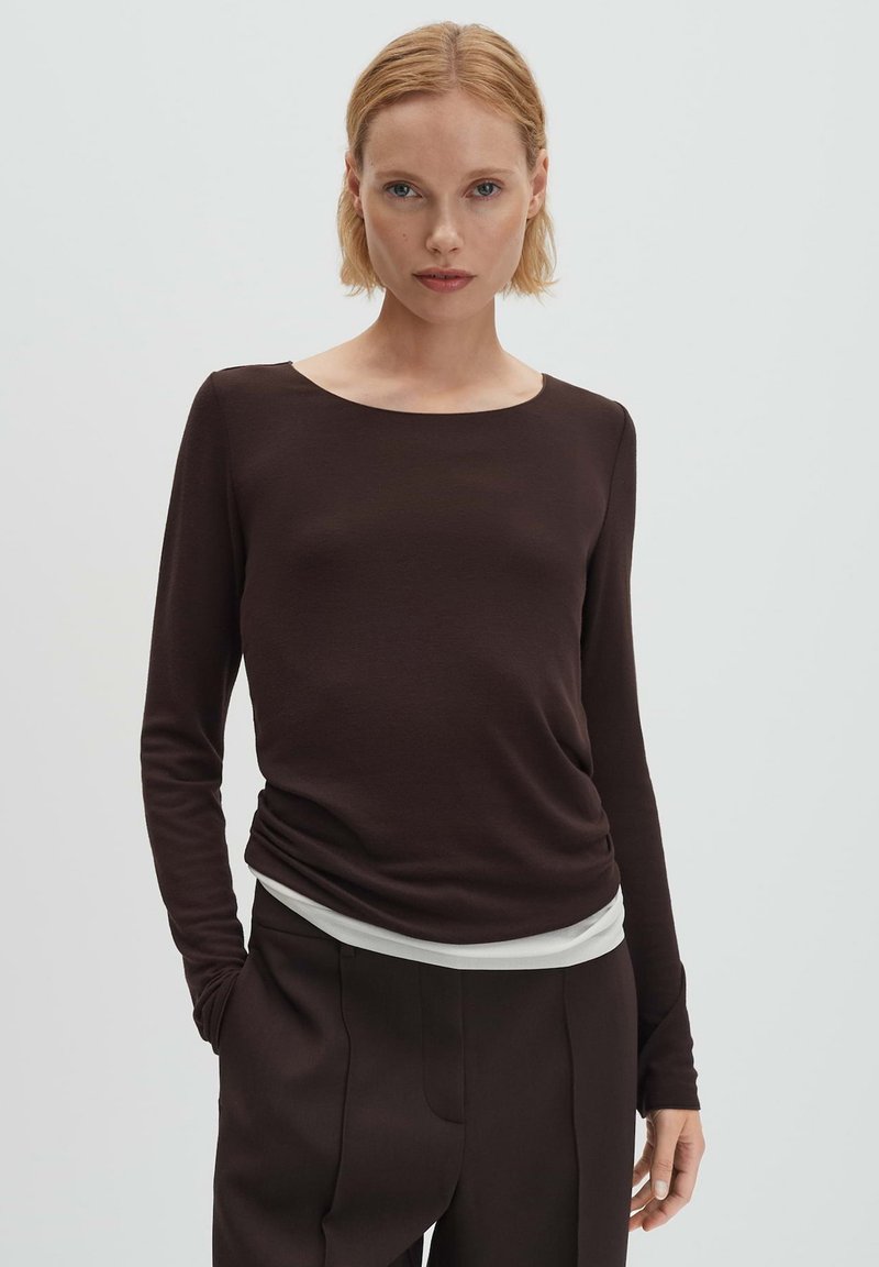 Braunfarbenes Langarmoberteil mit Rundhalsausschnitt, aus glattem Stoff, kombiniert mit einer hellen Basisschicht, die am Saum sichtbar ist.