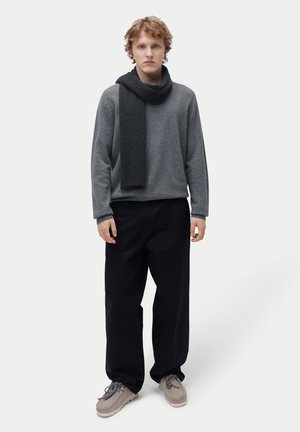 Junger Mann stehend, trägt einen grauen Pullover, dunklen Schal, schwarze weite Hose und beige Schuhe vor einem einfarbigen hellen Hintergrund.