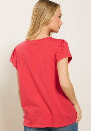 Vrouw met lang blond haar, gekleed in een rood kort mouwtopje en een blauwe spijkerbroek, van achteren gezien tegen een effen beige achtergrond.