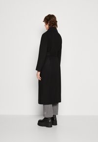 Sort lang frakke med et omslagssnor, der har en høj krave og lange ærmer. Modellen bærer grå cropped bukser og kraftige sorte støvler.