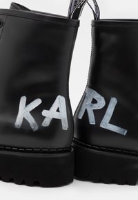 Botas de tobillo de cuero negro con letras pintadas en blanco que dicen "KARL", con una suela de goma texturizada y tiras laterales para facilitar el calzado.