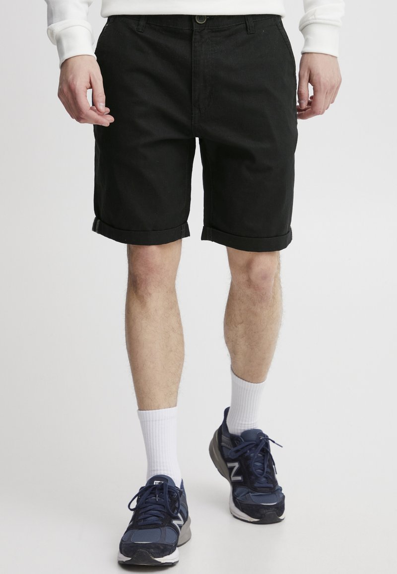 Shorts en denim noir avec une coupe classique, ourlets retroussés et détails de couture subtils, associés à des chaussures de sport navy et des chaussettes blanches.