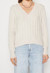 Pull beige en maille côtelée avec un col en V et des poignets ajustés ; associé à un jean en denim bleu clair. Le design met en avant la texture et le confort.