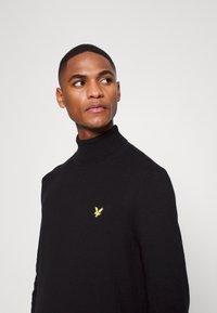 Maglione a collo alto di colore nero in morbido tessuto lavorato a maglia, con un piccolo logo giallo sul petto. Il design presenta una silhouette aderente e polsini a coste.
