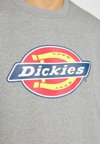 Camisa gris con un logo central de Dickies que presenta un círculo rojo, un herradura amarilla y una banda azul con texto blanco.