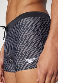 Sorte og grå mønstrede badetights med svart strikk i livet, med snor og en hvit Speedo-logo på siden. Glatt tekstur.