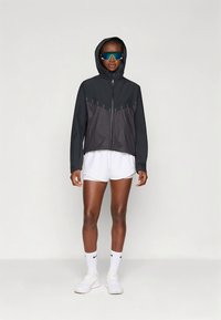 Svart och grå zip-up jacka med huva, med ett geometriskt mönster och reflekterande detaljer, bärs över vita shorts och träningsskor.