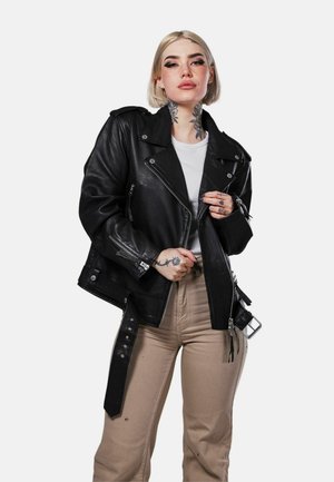 Jeune femme aux cheveux blonds courts, avec des tatouages floraux au cou et des tatouages aux mains, portant une veste en cuir noire, une chemise blanche et un pantalon beige.
