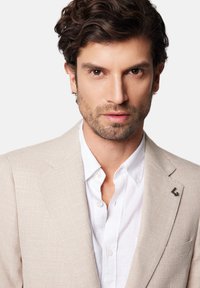 Jasno beżowa marynarka o teksturowanym wykończeniu, zestawiona z białą koszulą. Na klapie marynarki znajduje się małe ciemne logo.