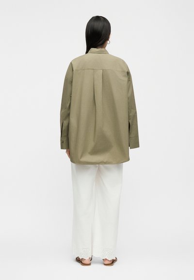 Γυναίκα με μακριά σκούρα μαλλιά που φοράει υπερμεγέθη πουκάμισο σε ελιά πράσινο, άσπρο παντελόνι με φαρδιά μπατζάκια και τελείωμα με τρύπες, και καφέ σανδάλια, με την πλάτη προς τα εμπρός.