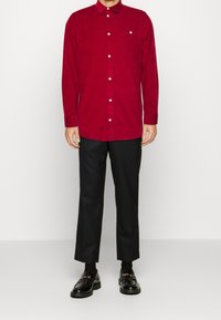Czerwona koszula z kołnierzykiem i małą kieszenią na piersi, w połączeniu z czarnymi spodniami i butami z dodatkami złotego okucia. Gładka tekstura.