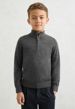 Grijze trui met ritssluiting, opgehoogde kraag en geribde boorden, gemaakt van textuurgebreide stof, gedragen met donkere broek. Minimalistische details overal.